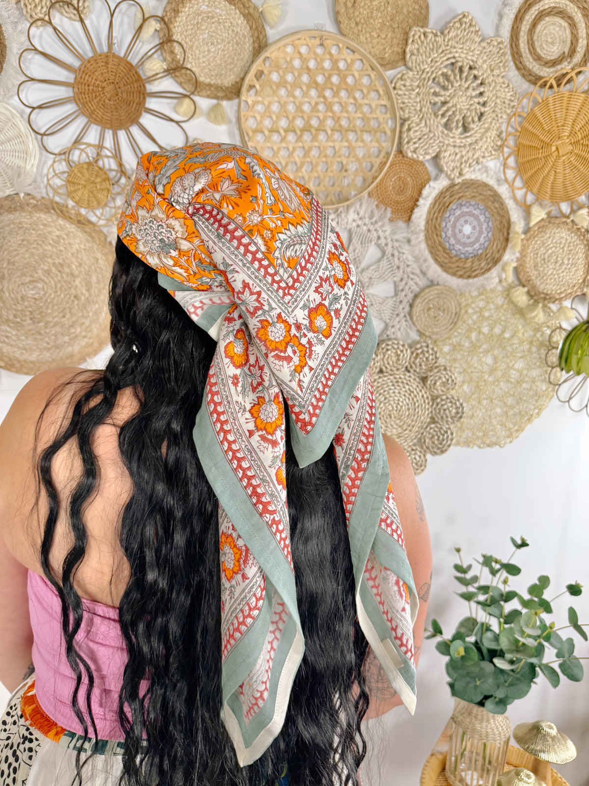 A woman with long, wavy black hair is shown from behind wearing a patterned scarf wrapped around her head and shoulders. The scarf features a vibrant orange and sage green bohemian-style print on a white background.