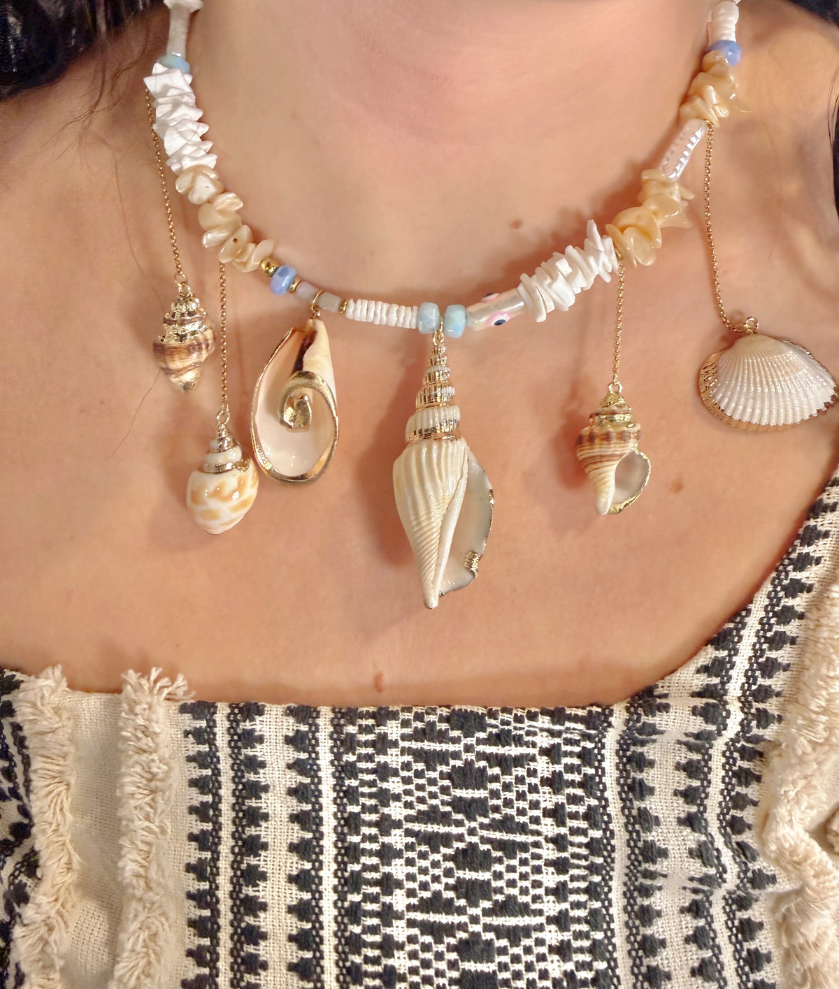 A close-up of a person wearing a bohemian-style seashell necklace with various natural conch shells and beads, paired with a black and white patterned top.