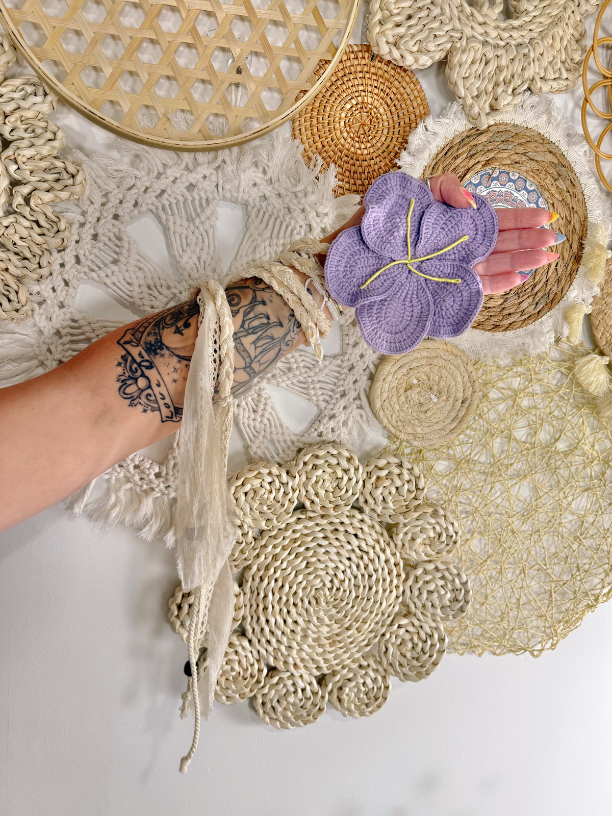 A hand with a sleeveless tattoo holds a lavender purple crocheted flower-shaped coaster with yellow stitching, surrounded by bohemian-style woven and crocheted decor on a white surface.