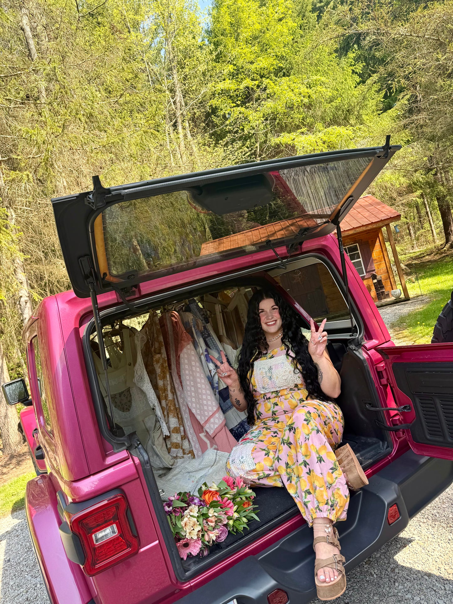 Woman sitting in the open trunk of a pink Jeep, surrounded by flowers and clothing, with a wooden cabin in the background.