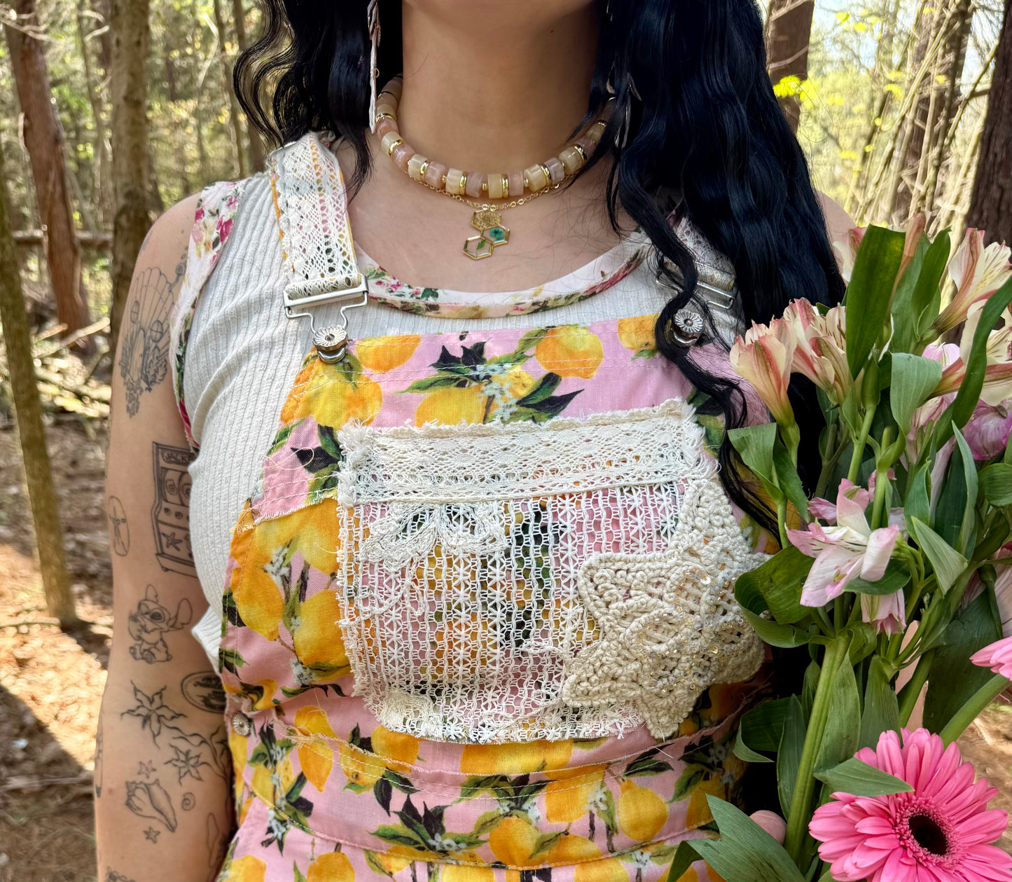 Person wearing a floral overalls with lace details, holding flowers in a forest setting