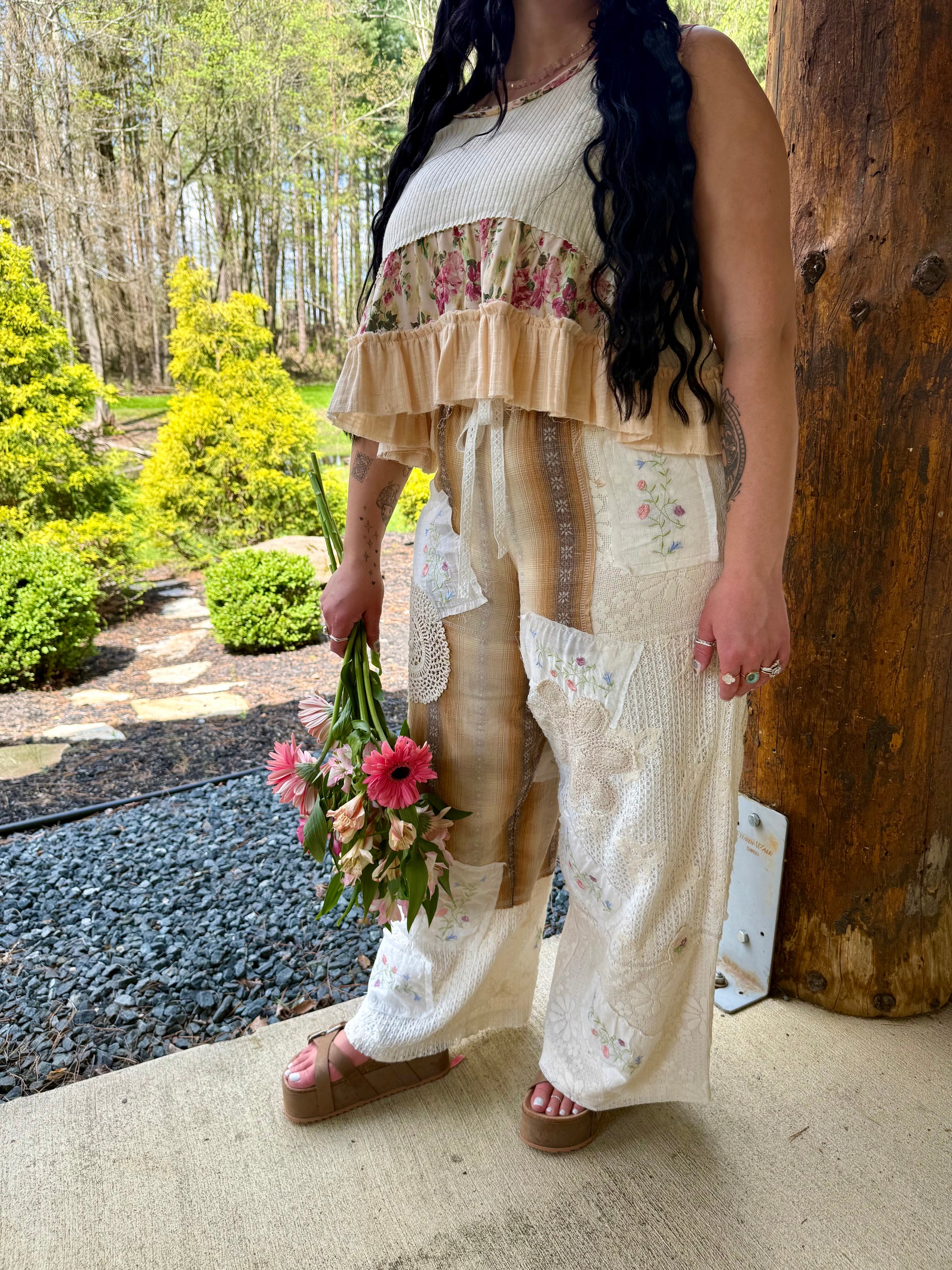 Woman in a floral top and beige pants holding flowers outdoors.