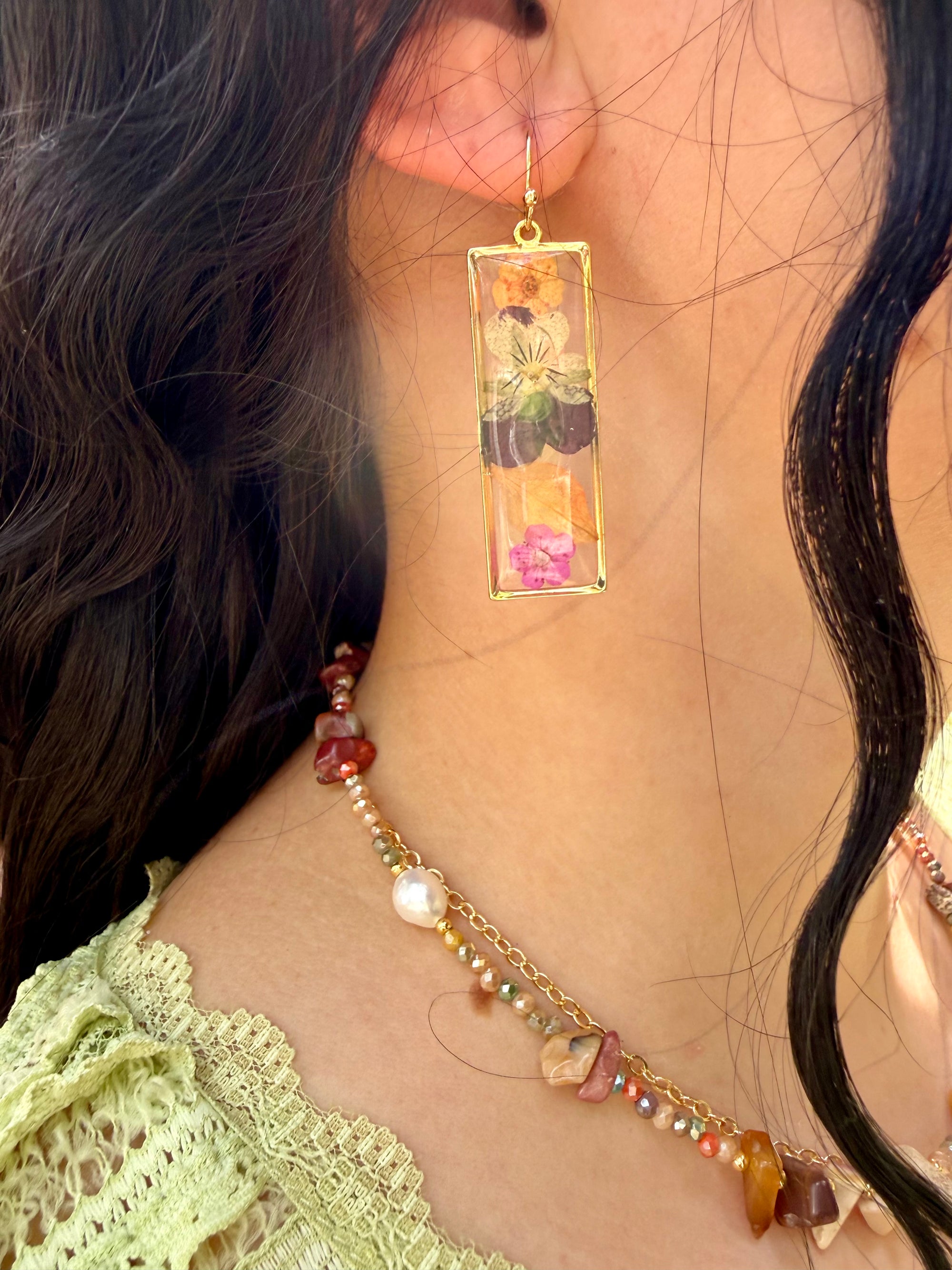 Close-up of a person wearing a floral rectangular earring and a beaded necklace.