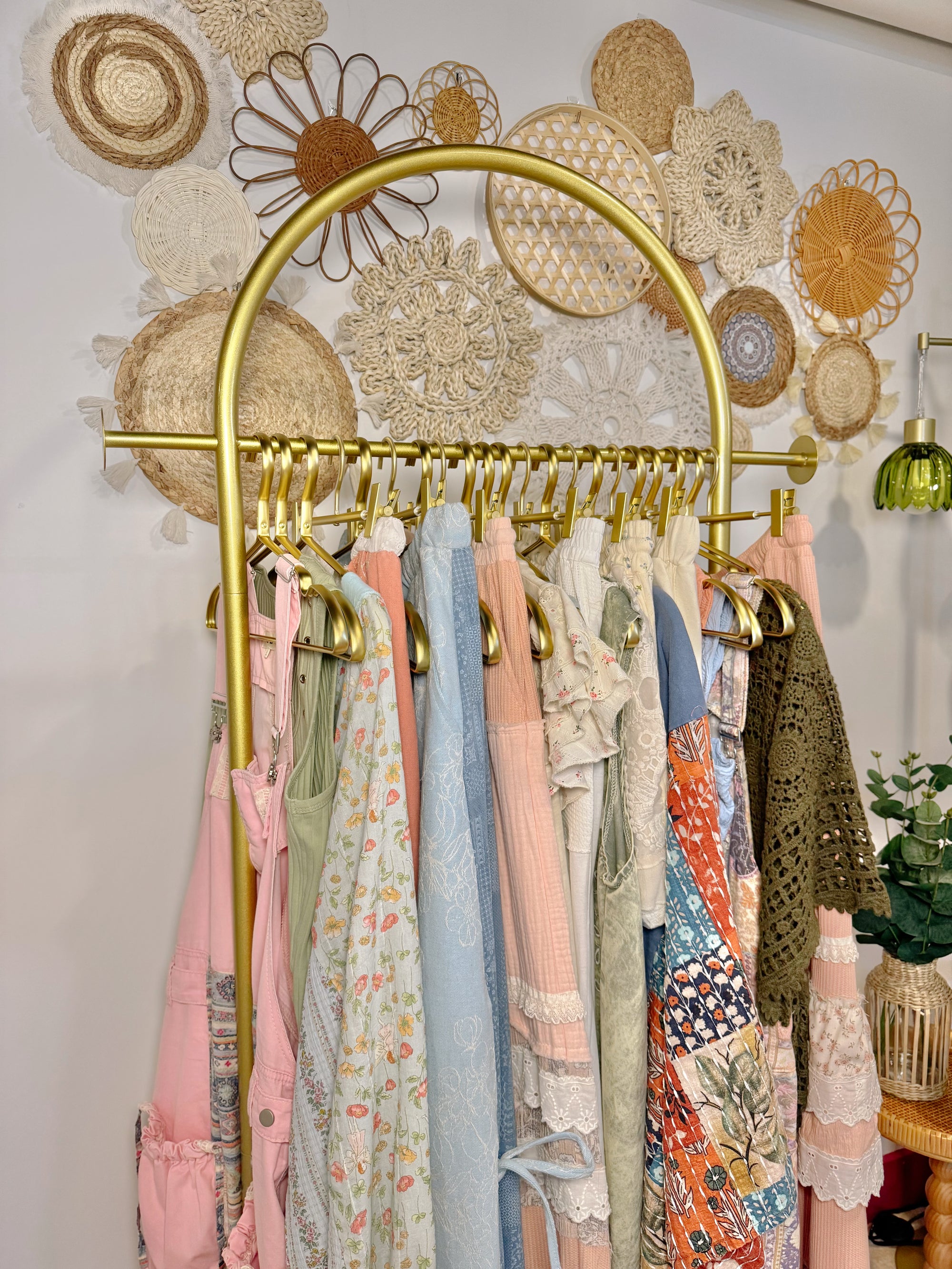A gold-framed clothing rack holding multiple pastel and patterned garments, set against a white wall decorated with woven wall art and a green hanging lamp.