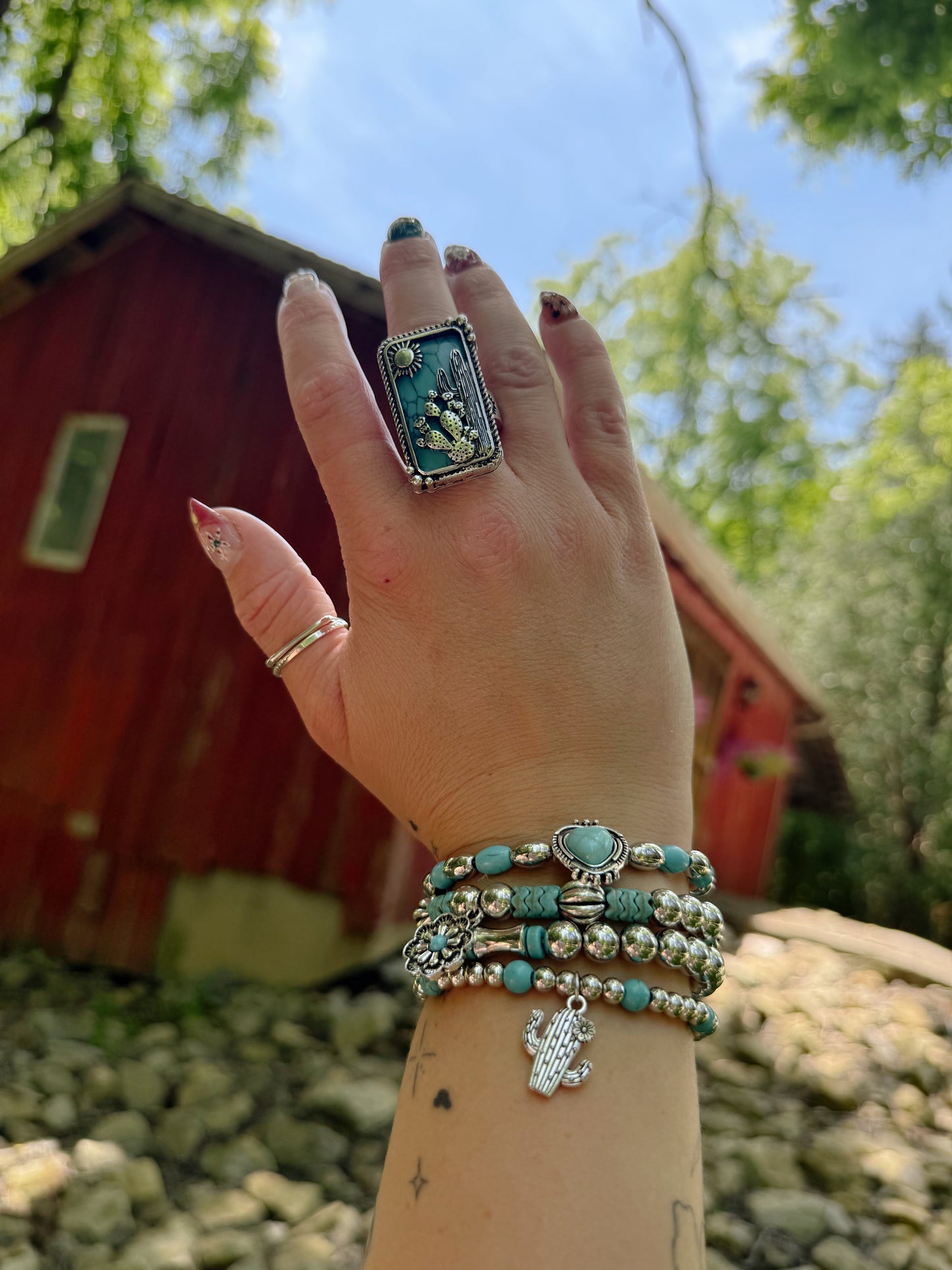 Hand wearing multiple bracelets and rings with a blurred background of a red building and greenery