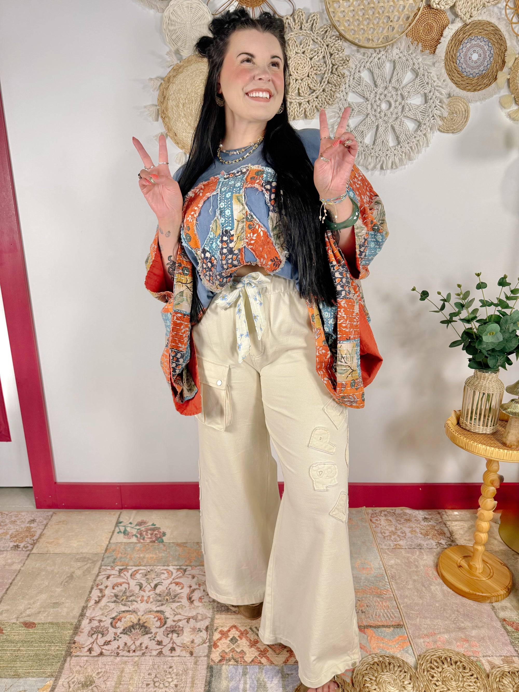 A woman posing in a bohemian-style outfit featuring a blue patterned top, an orange and blue patterned cardigan, and cream wide-leg pants, standing in front of a decorated wall.