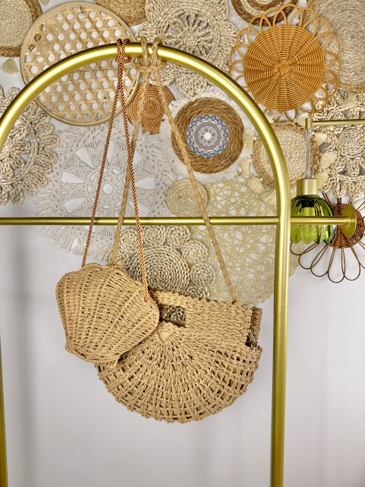 A woven straw shoulder bag with a matching small pouch hangs from a gold metal display stand against a white wall decorated with boho-style woven wall art.