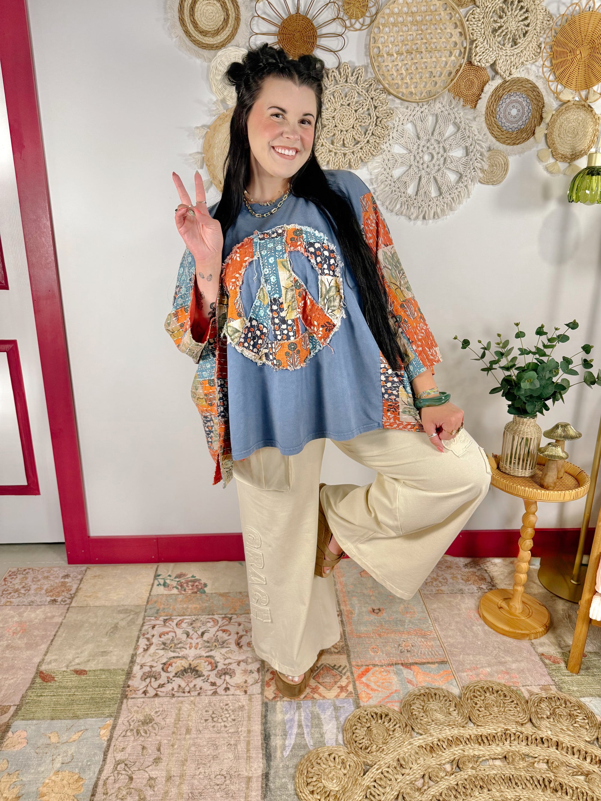 A woman posing in a bohemian-style outfit featuring a blue top with a colorful paisley peace sign graphic and patterned sleeves, paired with cream wide-leg pants, set against a decorated wall with woven wall art.