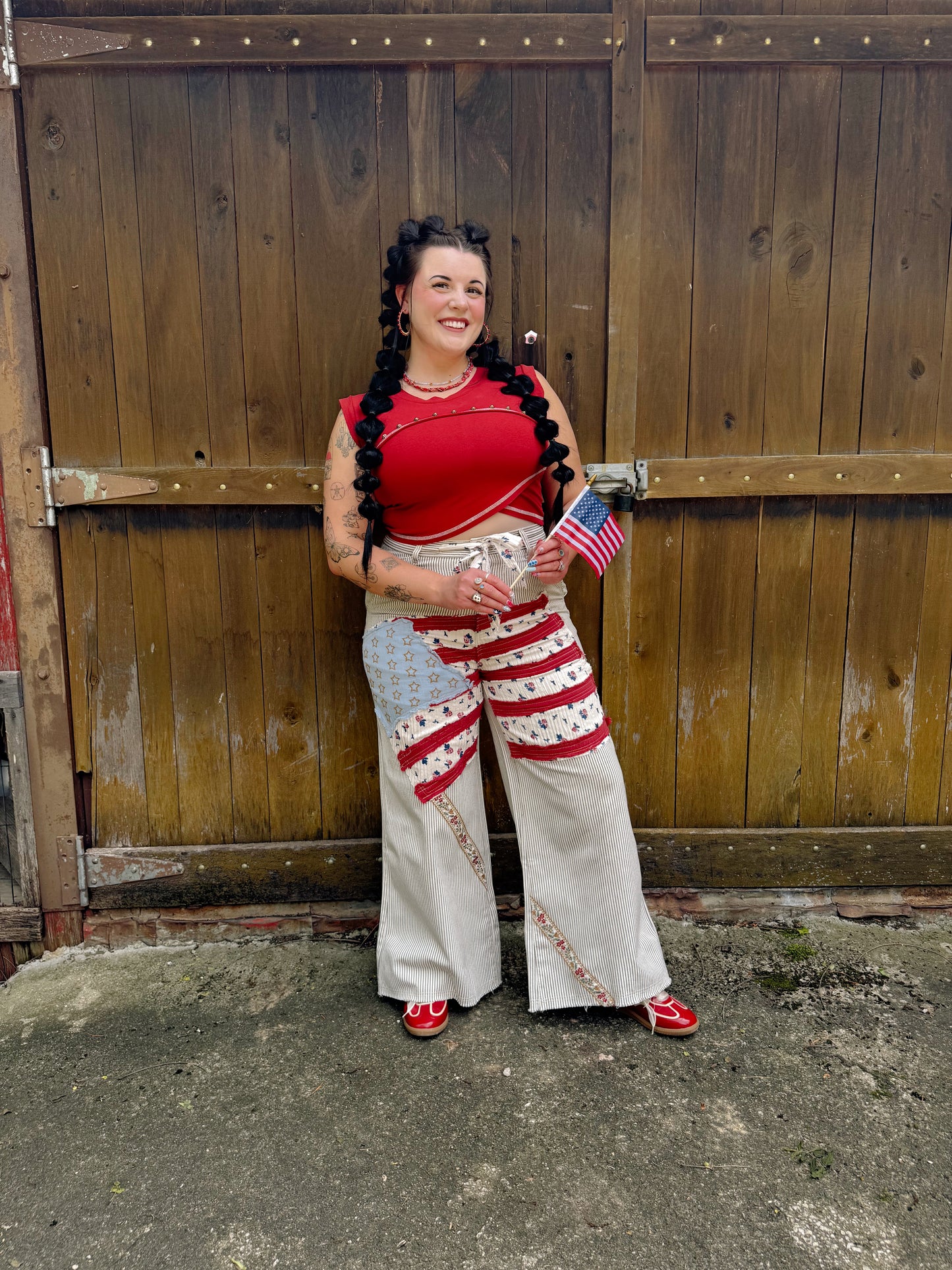 Woman in a red top and white pants with red and blue patterns standing in front of wooden doors.