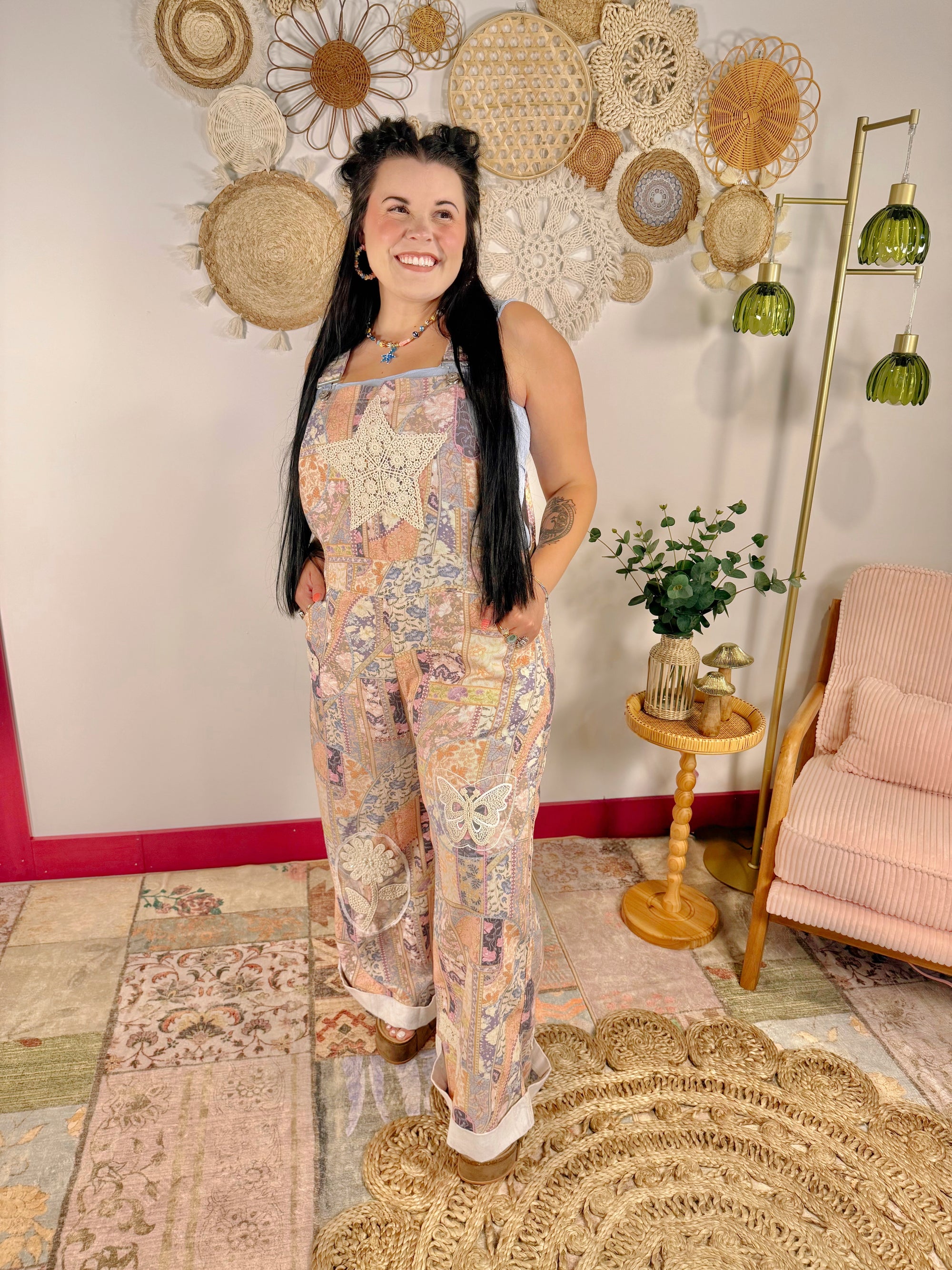 A woman stands indoors modeling a sleeveless, wide-leg jumpsuit with a pastel bohemian floral and geometric print. The background features a decorative wall with woven and crocheted elements, a gold floor lamp, and a pink armchair.