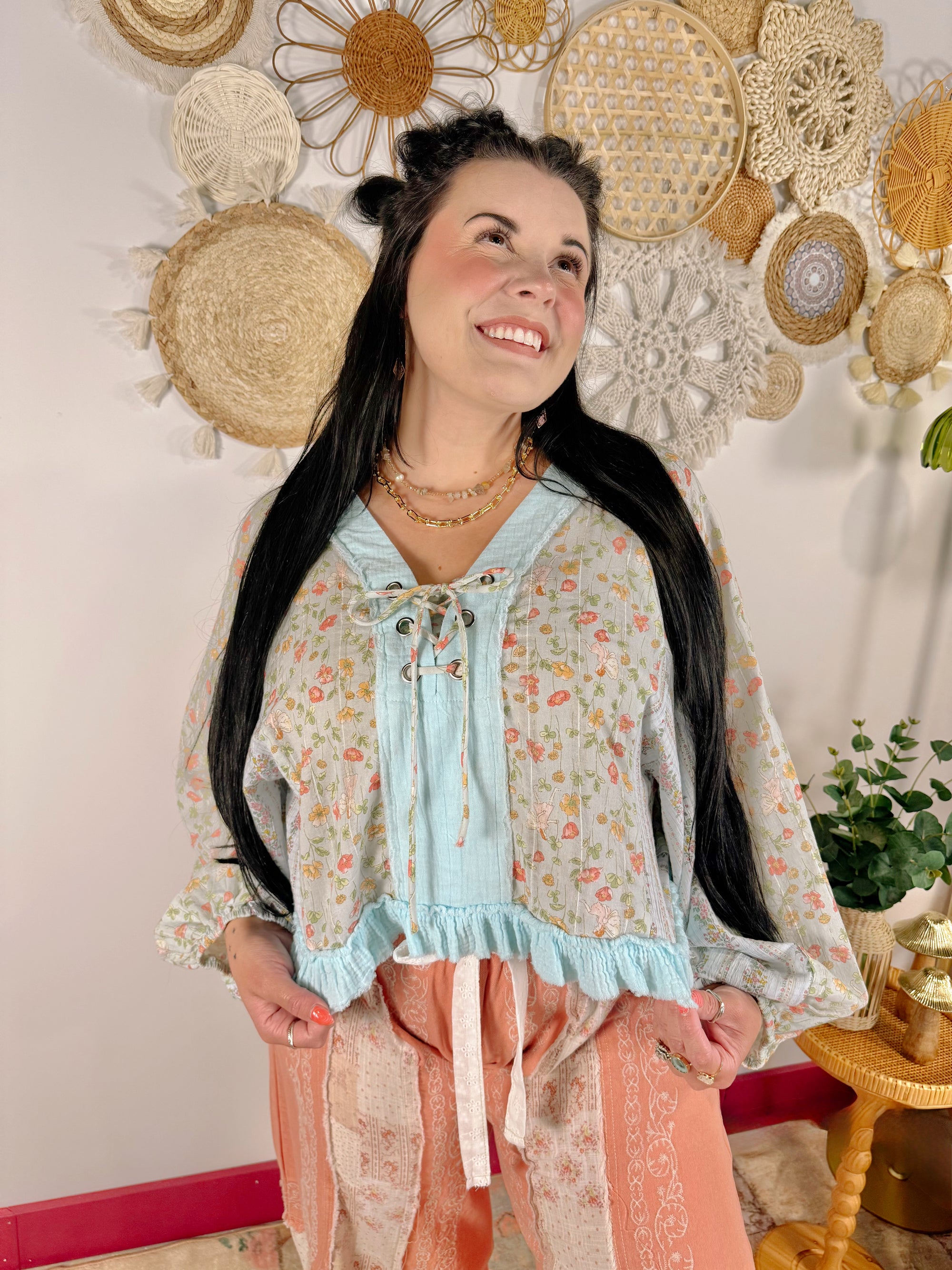 A woman with long black hair wearing a light blue and pink floral long-sleeve blouse with a tie-neck and ruffled hem, paired with peach patterned pants, standing in front of a wall decorated with woven wall art.