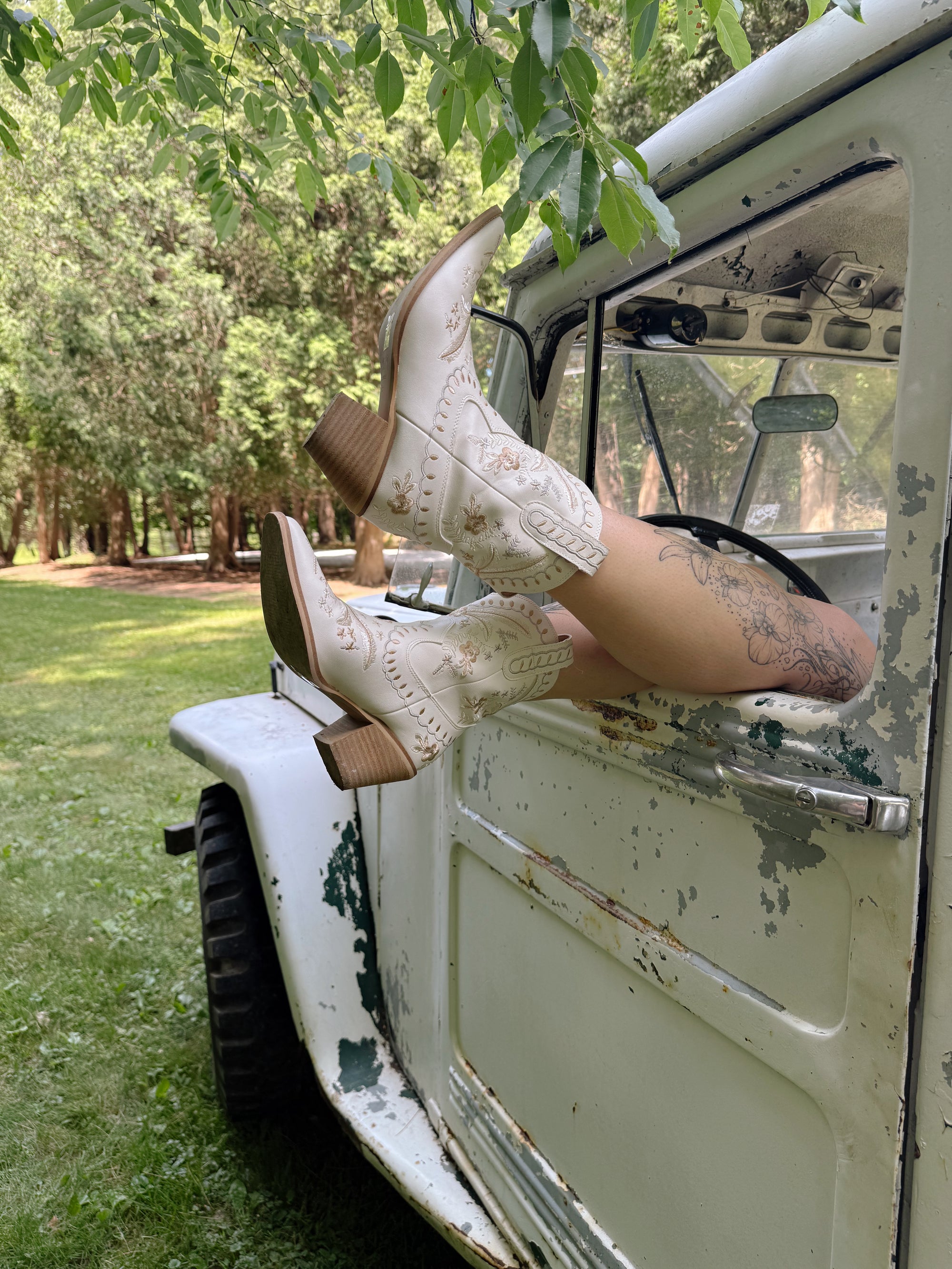 Person wearing white cowboy boots stepping out of a vintage vehicle with greenery in the background